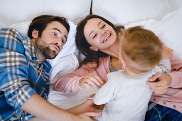 Parenthood concept. Caucasian young family of three parents mother and father with small little kid child toddler relaxing resting on the bed, playing and spending time together, taking care of kid