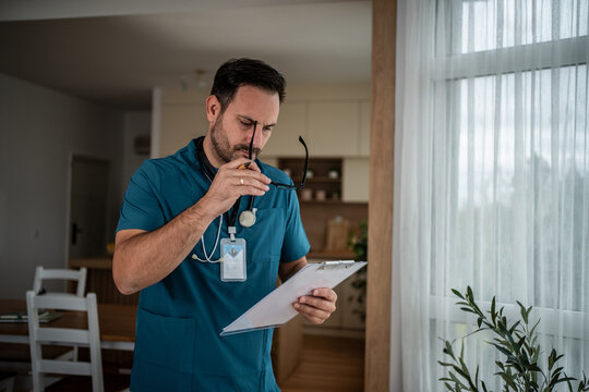 Male doctor holding glasses reviewing patient records during home visit - Powered by Adobe