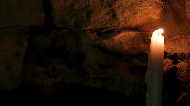 A single lit candle burns softly against an old textured stone wall in low light. Atmospheric video symbolizing solitude, silence, memory, or spirituality