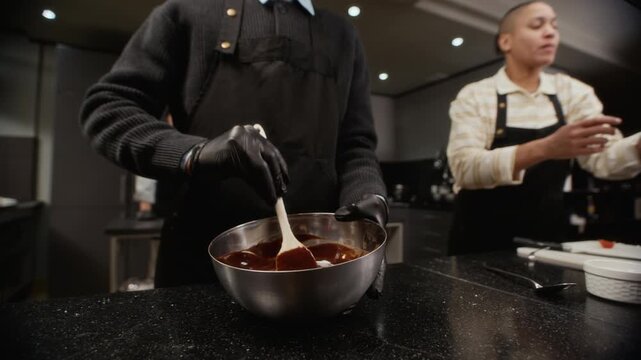 Gloved hands of culinary course trainee stirring melted chocolate with spatula in mixing bowl while cooking dessert during group class in restaurant kitchen