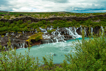 Hraunfossar is a unique Icelandic waterfall in West Iceland where clear, cold spring water seeps directly from under the Hallmundarhraun lava field, forming hundreds of small, delicate waterfalls.