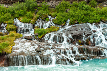 Obraz premium Hraunfossar is a unique Icelandic waterfall in West Iceland where clear, cold spring water seeps directly from under the Hallmundarhraun lava field, forming hundreds of small, delicate waterfalls.