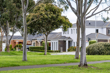 Leafy suburban street in Point Cook, Melbourne, Australia, featuring modern detached houses set behind a public green space with mature eucalyptus trees, grassed lawns, and pedestrian paths