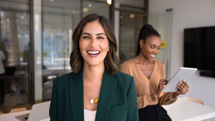 Obraz premium Happy beautiful Hispanic businesswoman posing for head shot in office workspace with diverse colleagues working behind, looking at camera, smiling. Banner portrait of confident female business leader