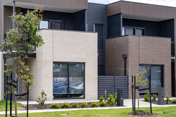 Modern low-rise townhouses in a newly developed suburban neighborhood in Australia, featuring small landscaped front yards with young trees. Contemporary medium-density housing design