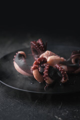 Obraz premium Hot prepared squid or boiled octopus with tentacles wiggling on a plate with steam coming out against black background. Seafood background for a restaurant menu