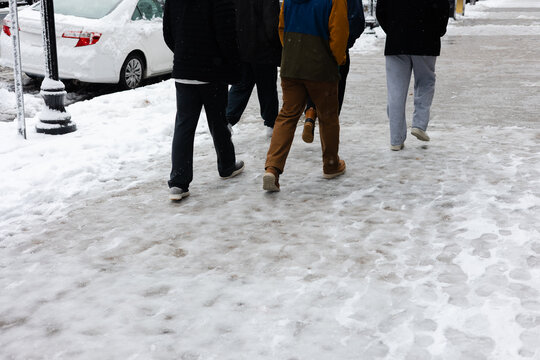 People walk on a snowy sidewalk in winter while snow falls lightly around them in a city setting