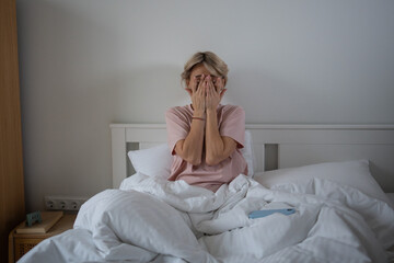 Tired and exhausted middle aged woman sitting in bed in bedroom feeling unwell, rubbing eyes...