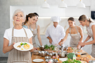 Elderly woman poses with prepared chicken dish at cooking master class