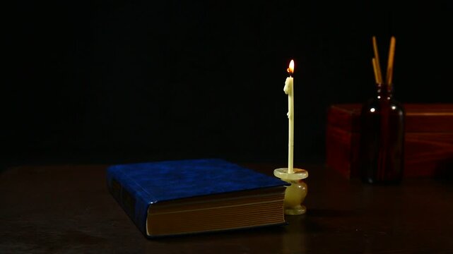 Burning wax white candle with spell book on the wood table, wooden box and bag on dark black background with empty copy space. Ancient old magic objects