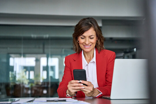 40s middle aged business woman holding mobile cell phone cellphone in office. Focused mature professional ceo entrepreneur businesswoman using smartphone for work sitting at desk workplace. Copy space