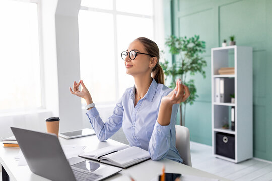 After a long day, a young female entrepreneur sits at her desk in an office, eyes closed, practicing meditation and deep breathing exercises to manage work stress and find calmness.
