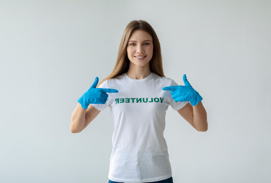A young caucasian woman wears a volunteer t-shirt and blue gloves, smiling warmly as she points to herself. She stands in front of a light wall, showcasing her pride in helping at a charity. - Powered by Adobe