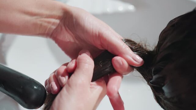 close-up of stylist twisting and wringing wet hair to remove excess water at salon sink, careful handwork preps hair for blowdry or cut with clinical white basin and focused technique