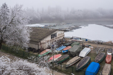 Foggy winter boat harbor with frozen water &ndash; Small boats stored near icy lake, misty winter landscape with frost covered trees and calm atmosphere.