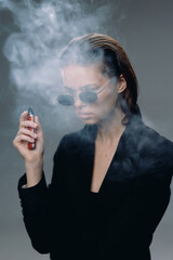 Woman vaping with sunglasses and black jacket surrounded by vapor smoke in dark setting