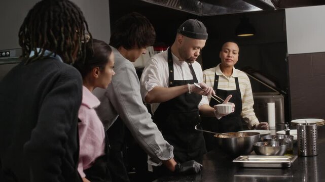 Male chef holding ramekin topping dish using plating tweezers while giving tutorial to multiethnic group of young trainees at restaurant kitchen workbench