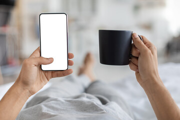 Male hands are relaxed in bed, holding a smartphone with a blank screen in one hand and a cup of coffee in the other. A cozy home setting is visible in the background, offering a moment of leisure.