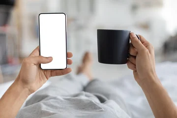 Fotobehang Muziek Male hands are relaxed in bed, holding a smartphone with a blank screen in one hand and a cup of coffee in the other. A cozy home setting is visible in the background, offering a moment of leisure.  © Prostock-studio