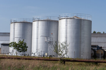 Three large tanks are in a field. The tanks are silver and are next to a fence. There is a tree in the background