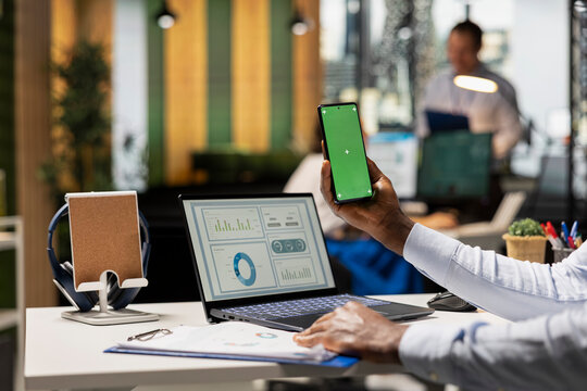 African american manager preparing company progress report, holding smartphone with green screen while reviewing financial charts on laptop. Male analyst using digital devices in modern workspace. - Powered by Adobe