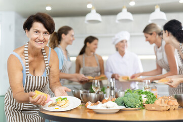 Elderly woman in apron pours sauce into ready dish at cooking master class