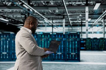 Admin walking in data center running lines of code on laptop, troubleshooting rackmounts. African american IT expert using programming scripts on notebook device in server room