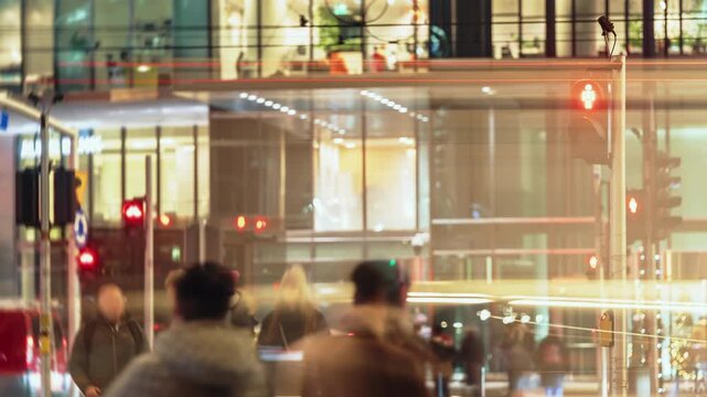 Timelapse of busy downtown city intersection at night with pedestrians walking crosswalk, traffic lights and blurred light trails from passing cars and trams. Modern architecture illuminated windows