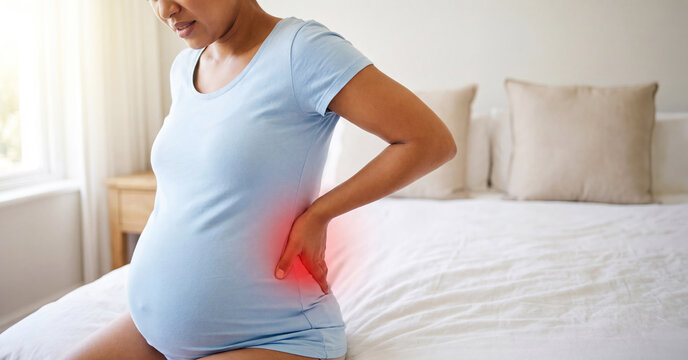 A pregnant woman is seated on her bed in a well-lit room during the day, gently holding her lower back as she relaxes - Powered by Adobe