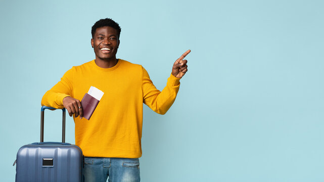 Smiling black man with plane tickets, passport and suitcase posing over blue studio background, pointing at copy space, panorama. Stylish african american guy travelling while COVID-19 pandemic