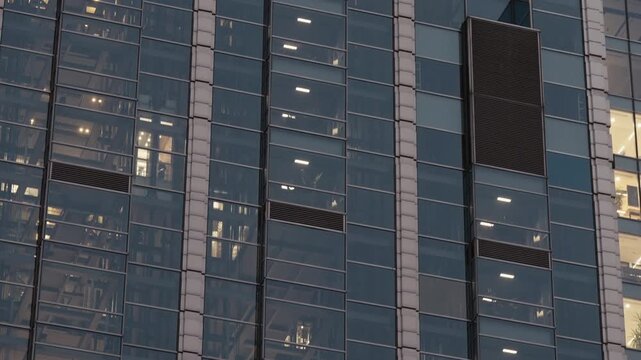 Modern office building in evening with glass elevators moving up and down behind illuminated windows. Urban architecture, business activity and rhythm of downtown life