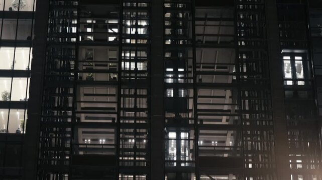 Modern office building in evening with glass elevators moving up and down behind illuminated windows. Urban architecture, business activity and rhythm of downtown life