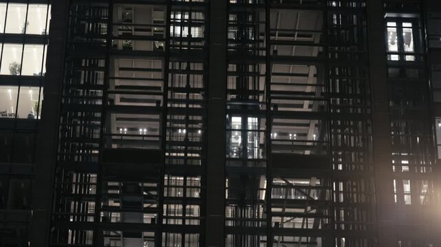 Modern office building in evening with glass elevators moving up and down behind illuminated windows. Urban architecture, business activity and rhythm of downtown life