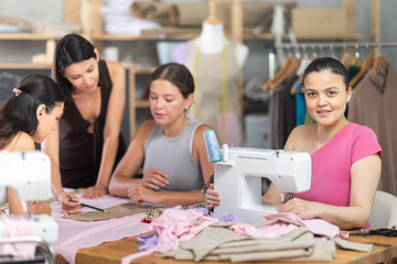 Interested young Asian woman working at sewing machine during hands-on course for adults, surrounded by classmates preparing patterns, fabric and fitting garments..