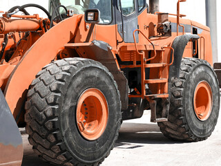 Side view of a heavy orange industrial front loader parked on a construction site. Focus on the large treaded tires, hydraulic lift arms, and metal access ladder