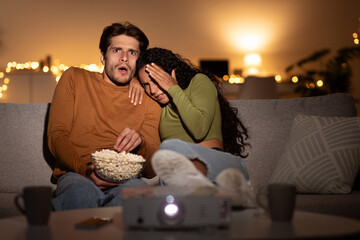 A couple sits on a sofa in their living room, shocked while watching a horror movie. They cover their eyes and share popcorn. The setting is cozy with dim lighting and home cinema vibes. © Prostock-studio