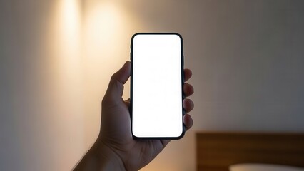 Hand holding smartphone with white screen in minimalist bedroom with soft lighting