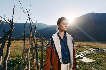 Naklejka premium Woman portrait in mountains with warm sunlight, fashion outfit and layered jacket standing outdoors by rustic wooden fence, serene landscape and travel lifestyle mood.