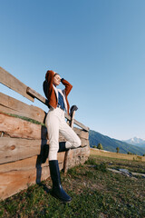 Fototapeta premium Woman leaning on wooden fence in mountains meadow, wearing fashion boots and casual outfit, outdoor countryside portrait with clear sky and relaxed travel lifestyle.