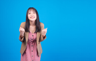 Excited young Asian woman business person celebrating success with raised fists on blue background. Happy female entrepreneur winning and cheering concept with copy space for text.