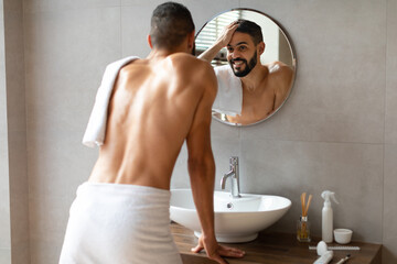 Young man smiles at his reflection in the mirror while touching his hair after a shower. He is...