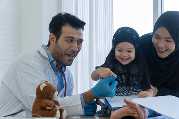 Friendly male doctor playing with cute Muslim little girl patient and mother in clinic. Happy healthcare professional interacting with kid near teddy bear for pediatric concept.