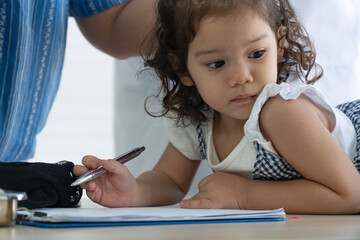Adorable little girl holding pen looking away while doing homework at home. Distracted cute female kid studying concept with parent standing nearby in background. (167 characters
