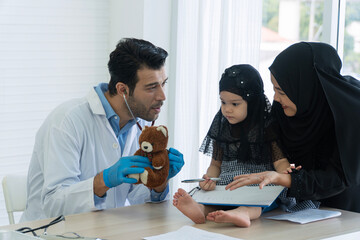 Friendly pediatrician doctor using stethoscope to check teddy bear near cute Muslim girl patient. Friendly medical checkup concept to build trust with kid in clinic.