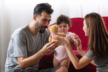 Happy multi-ethnic family playing with teddy bear at home. Middle Eastern father and Asian mother having fun with cute baby daughter in living room lifestyle concept.