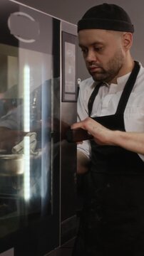 Vertical shot of adult male chef wearing apron taking tray of freshly baked chocolate cupcakes from oven using towel in restaurant kitchen
