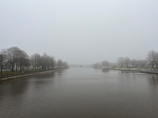 Fototapeta premium Foggy river scene with calm water, bare trees lining both banks, and a distant bridge barely visible through thick mist, creating a serene and atmospheric winter landscape.