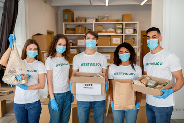 Fototapeta premium Five young volunteers pose indoors at a charity organization center. They are wearing gloves and masks while holding donation boxes filled with food items. Their teamwork promotes community support.