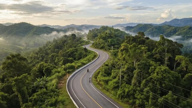 An breathtaking aerial view reveals a winding asphalt road cutting through lush, verdant mountains covered in dense forest. Mist gently clings to the valleys and distant peaks, creating a serene and m