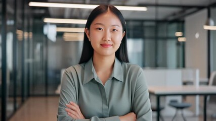 Confident young Asian businesswoman in a modern corporate office, standing with arms crossed and looking directly at the camera, embodying professionalism and determination.